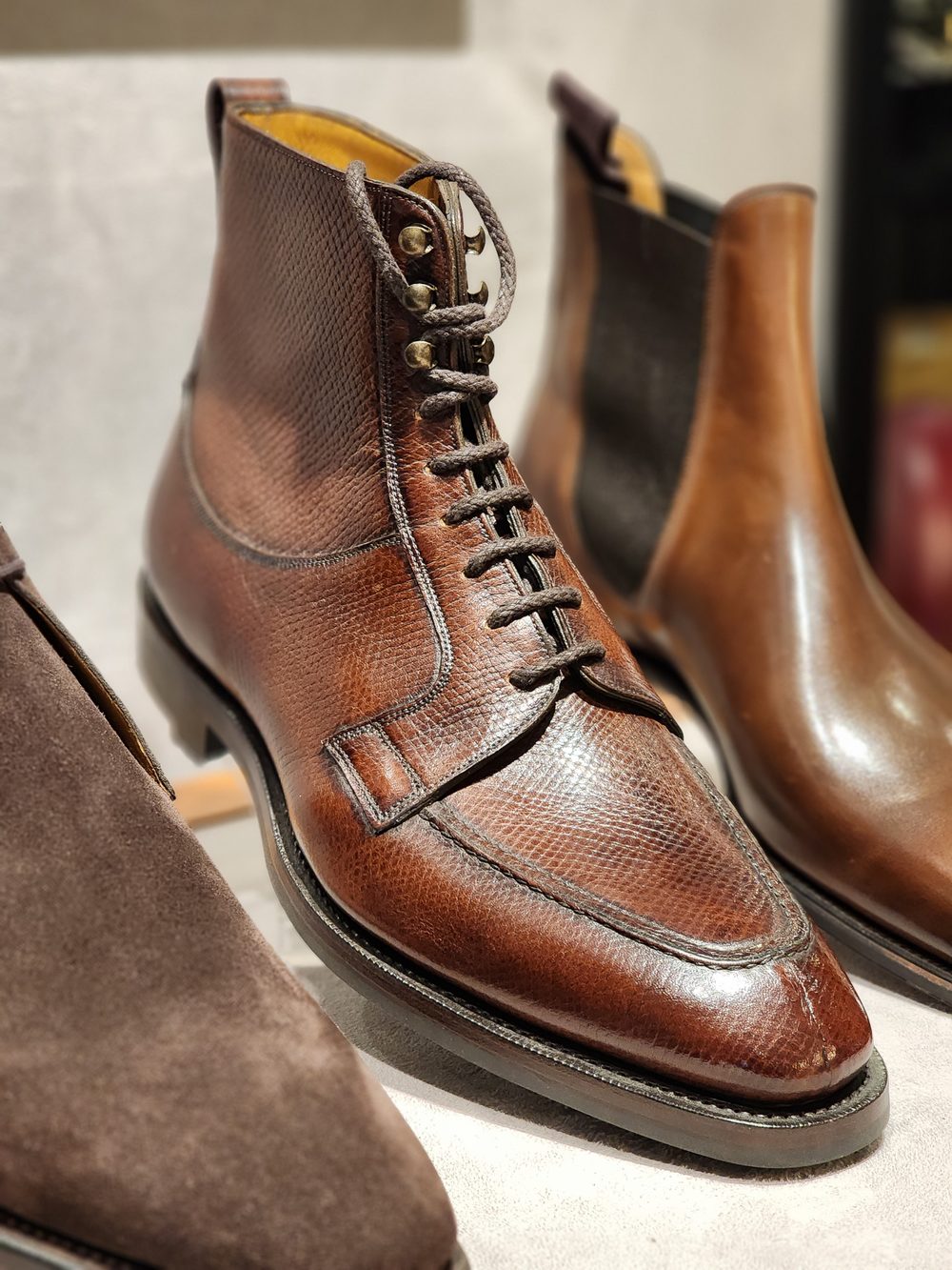 A close-up of a brown leather boot featuring a textured surface, lacing, and decorative eyelets, alongside two other shoes.
