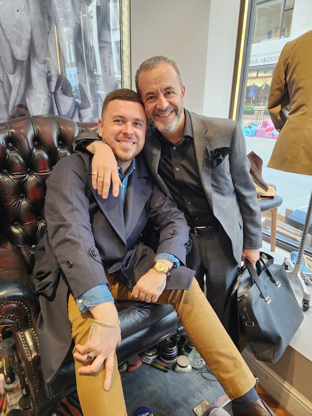 Two men smiling together, one in a navy coat and the other in a gray suit, seated on a leather chair.