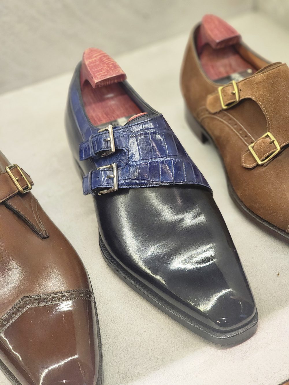 A polished black leather shoe with blue crocodile accents and double buckles, flanked by brown suede and brown leather shoes.