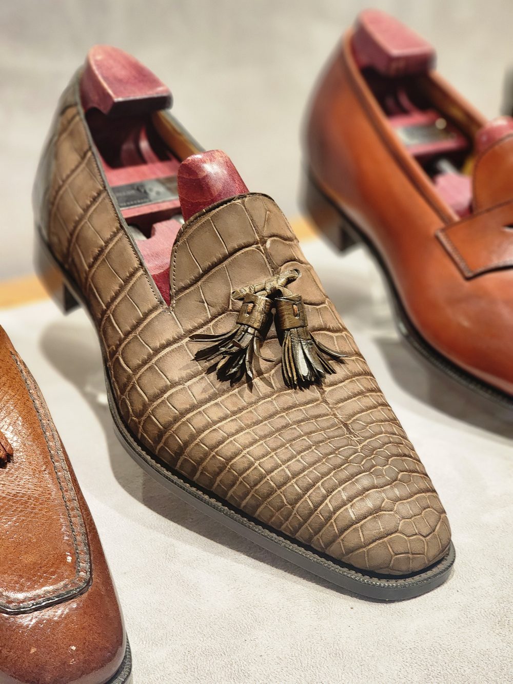 A brown crocodile leather loafer with tassels is displayed alongside other shoes.