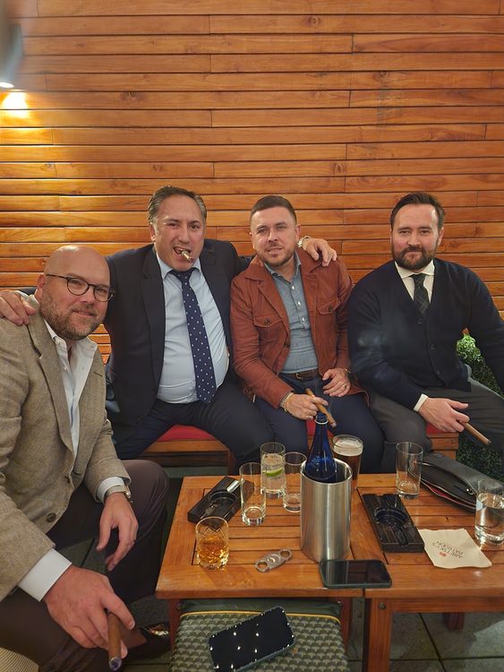 Four men are seated together, one holding a cigar, with drinks and a bottle on a wooden table in front of them.