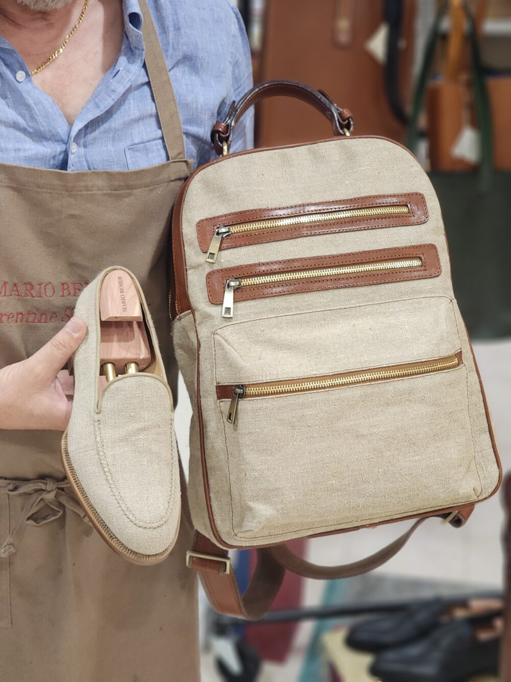 A person holds a pair of beige Mario Bemer canvas loafers in one hand and a matching beige backpack with brown leather accents in the other.
