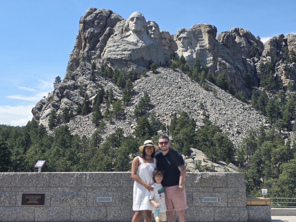 A family of three stands in front of Mount Rushmore, showcasing the carved faces of four U.S. presidents on the granite rock.