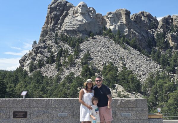 A family of three stands in front of Mount Rushmore, showcasing the carved faces of four U.S. presidents on the granite rock.