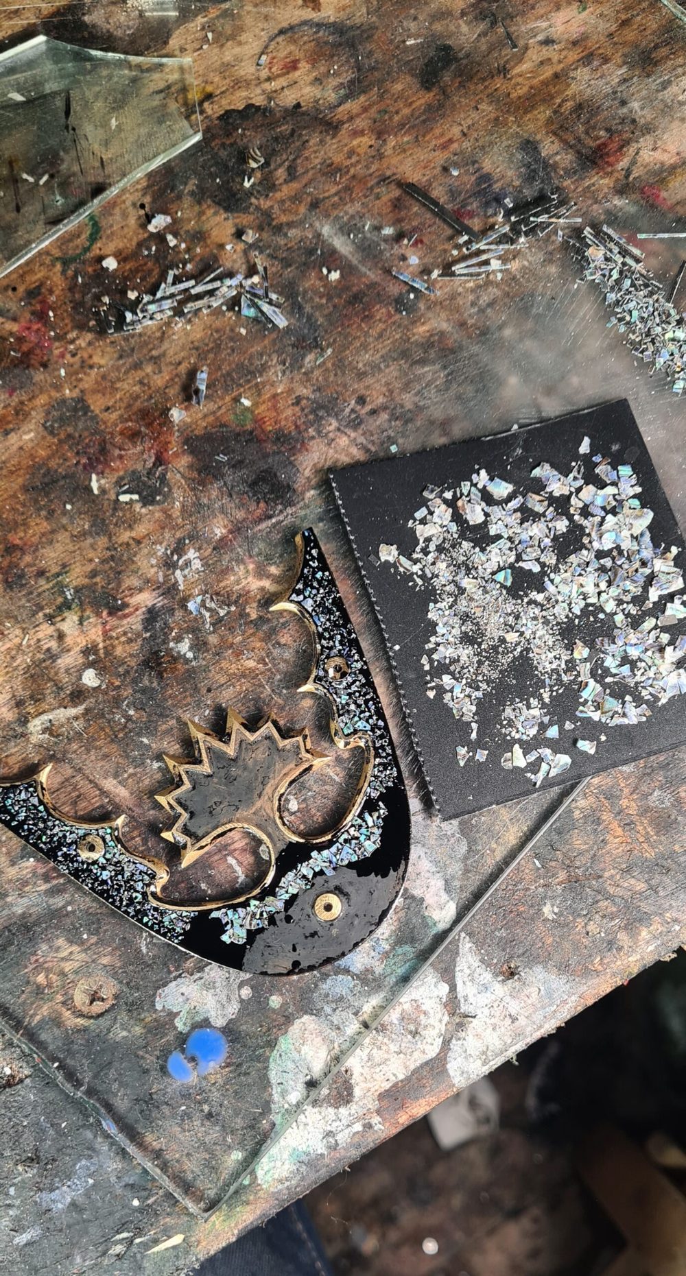 A black and gold shoe piece with decorative glitter is placed next to a pile of shiny, iridescent shards on a work surface.