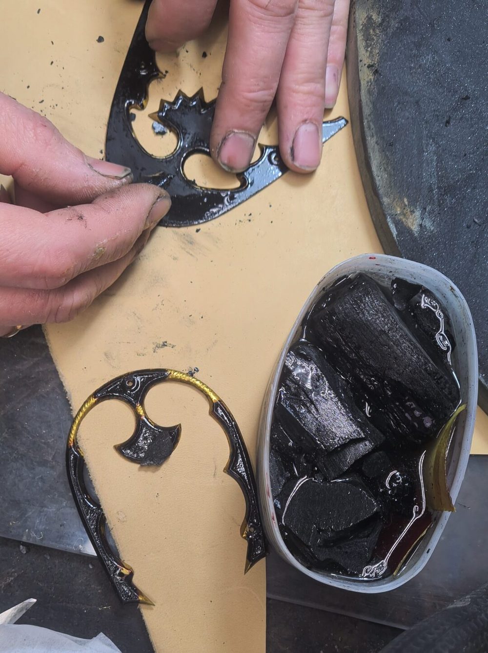 A close-up of a person's hands working on a black and gold decorative shoe component, with a container of black material nearby.