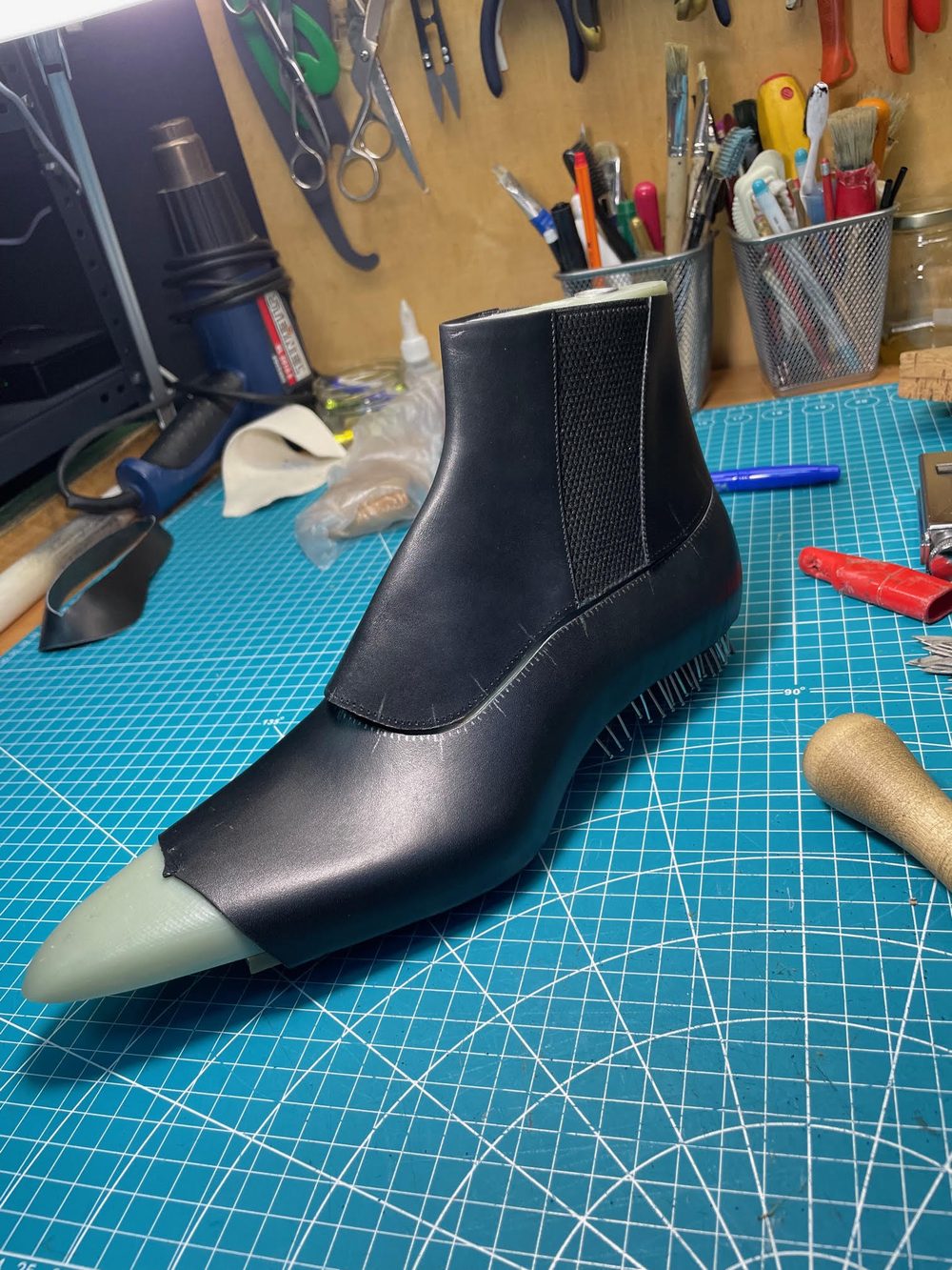 A black and green shoe prototype with a pointed toe and textured sides, displayed on a blue cutting mat.