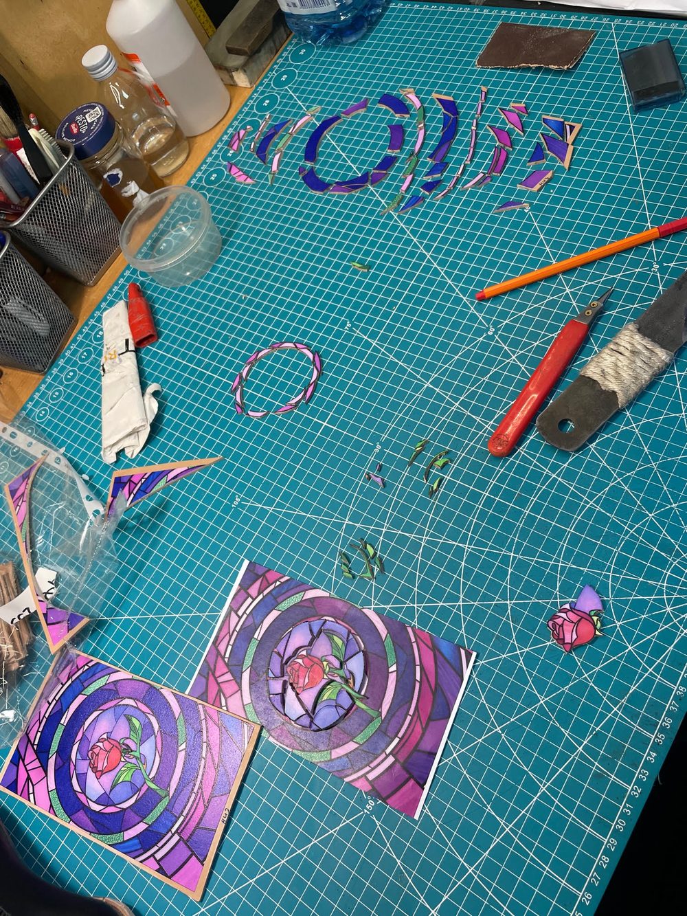 A workspace featuring colorful shoemaking materials, including purple and pink cutouts and a rose design on two cards.