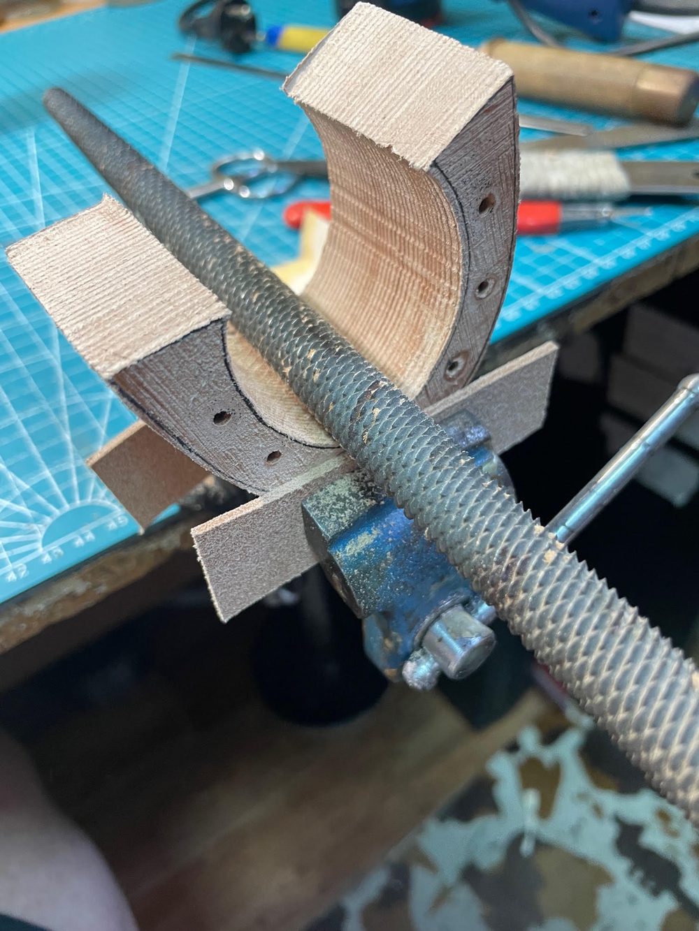 A wooden shoe last is held in a vise next to a metal rod, with various tools visible in the background.
