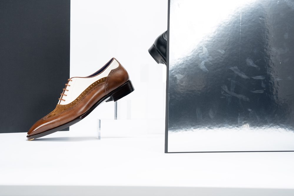 A brown and white dress shoe is displayed prominently, showcasing its sleek design and brogue detailing.