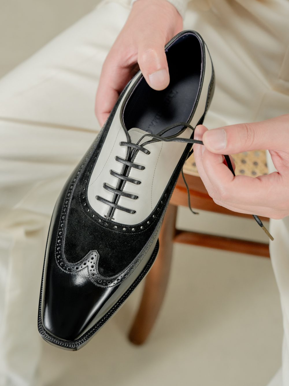 A hand is tying the laces of a two-tone black and white spectator shoe made of leather and suede.