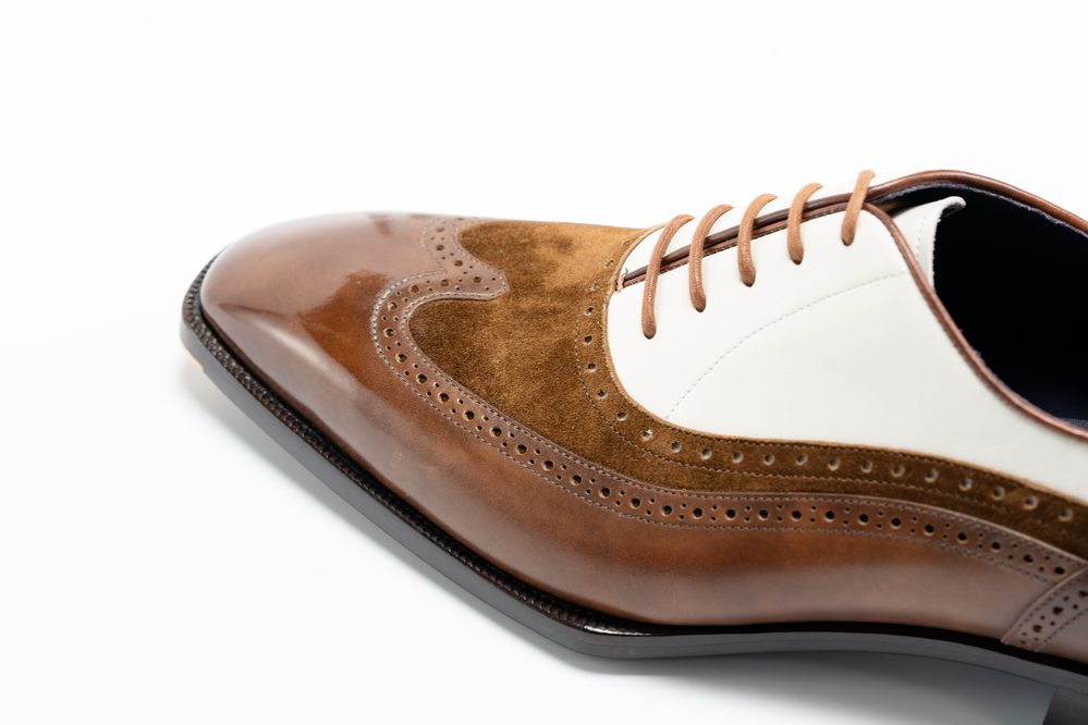 A close-up of a two-tone spectator shoe featuring a shiny brown leather toe and a white leather upper with brown suede accents.
