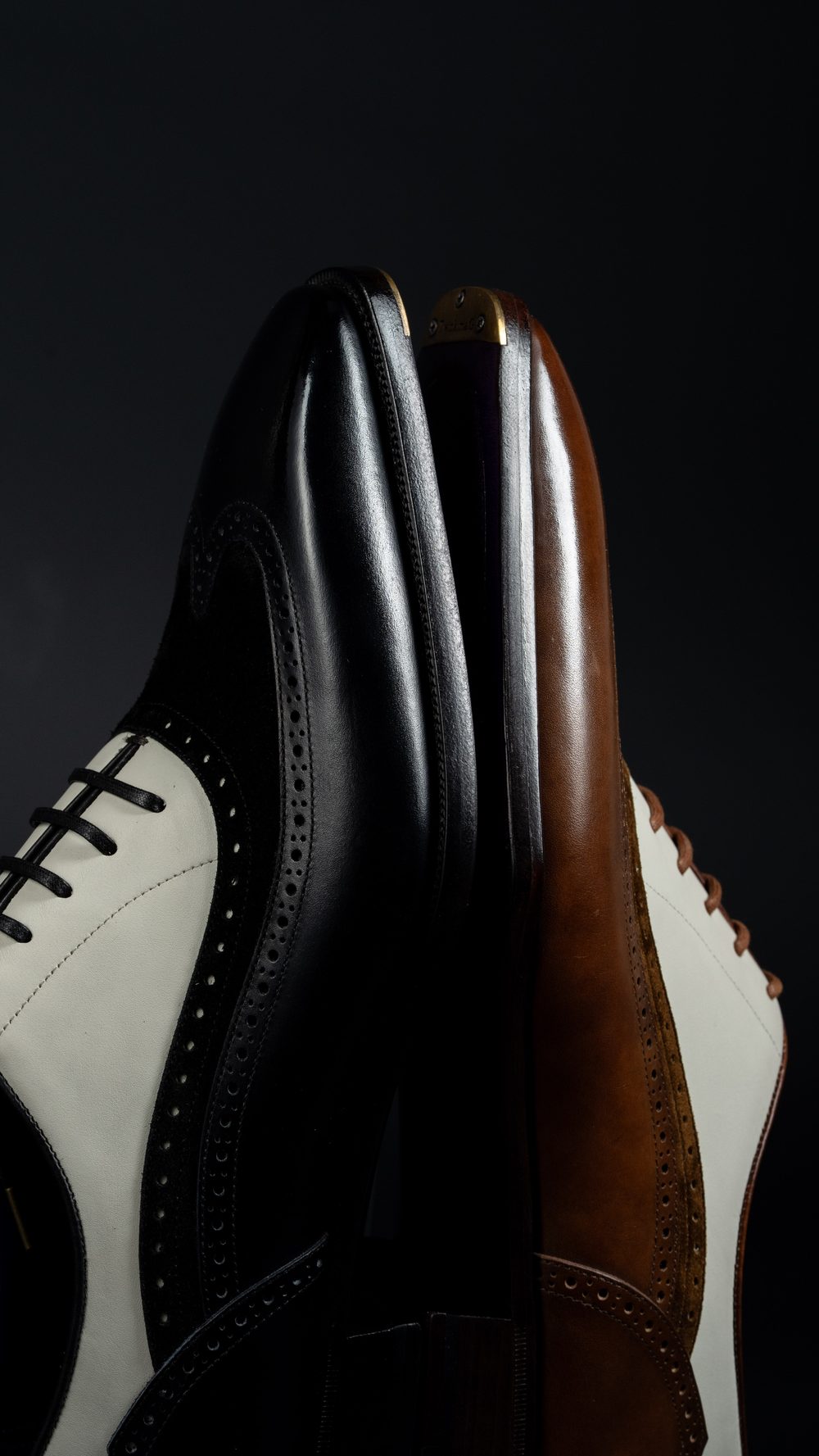 A close-up of two stylish spectator shoes, one black and white and the other brown and white, showcasing their sleek leather design.