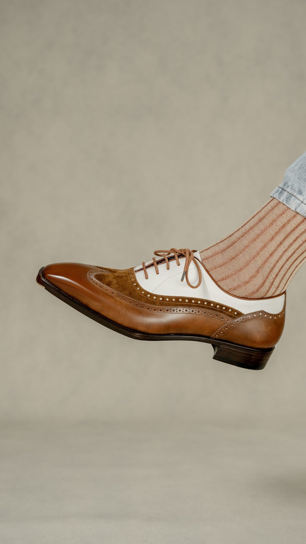 A brown and white spectator shoe is shown with a foot wearing a light striped sock.
