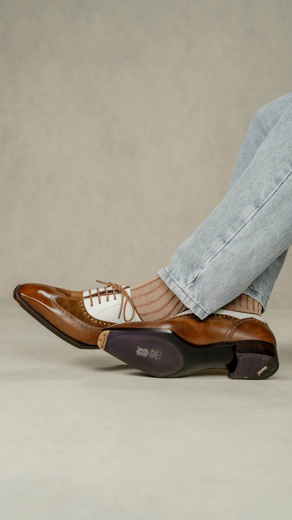 A side view of a brown and white spectator shoe paired with light-colored striped socks and denim jeans.
