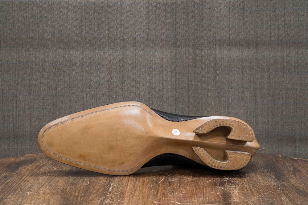 The image shows the sole of a black shoe with a tan leather bottom and a distinctive heel design.