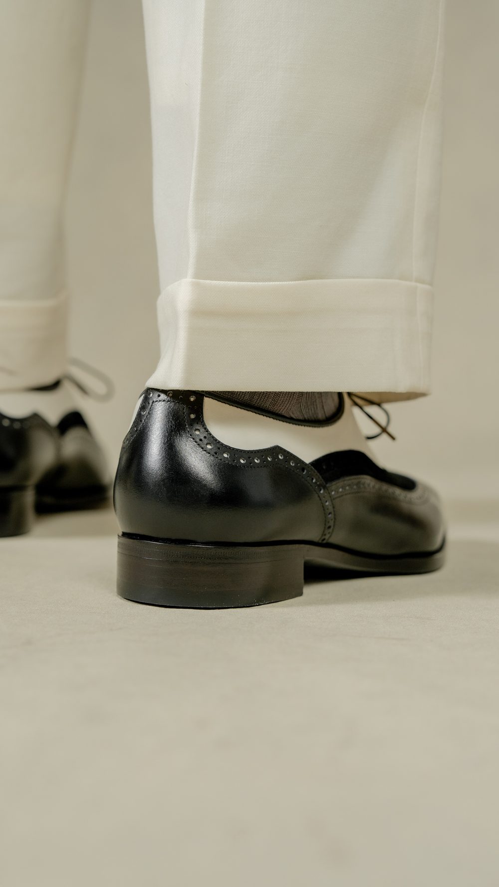 Close-up of a black leather spectator shoe with decorative perforations, paired with cream-colored trousers.