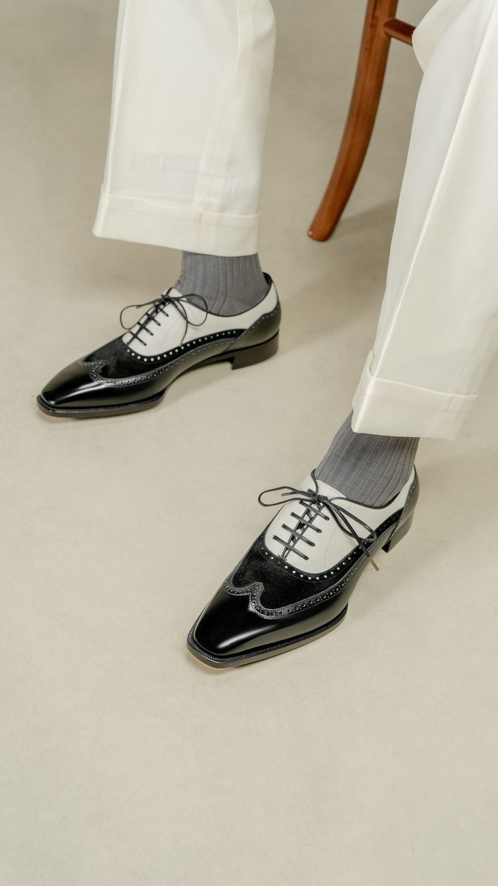 A close-up of black and white dress shoes with gray socks, worn by a person in cream-colored pants.