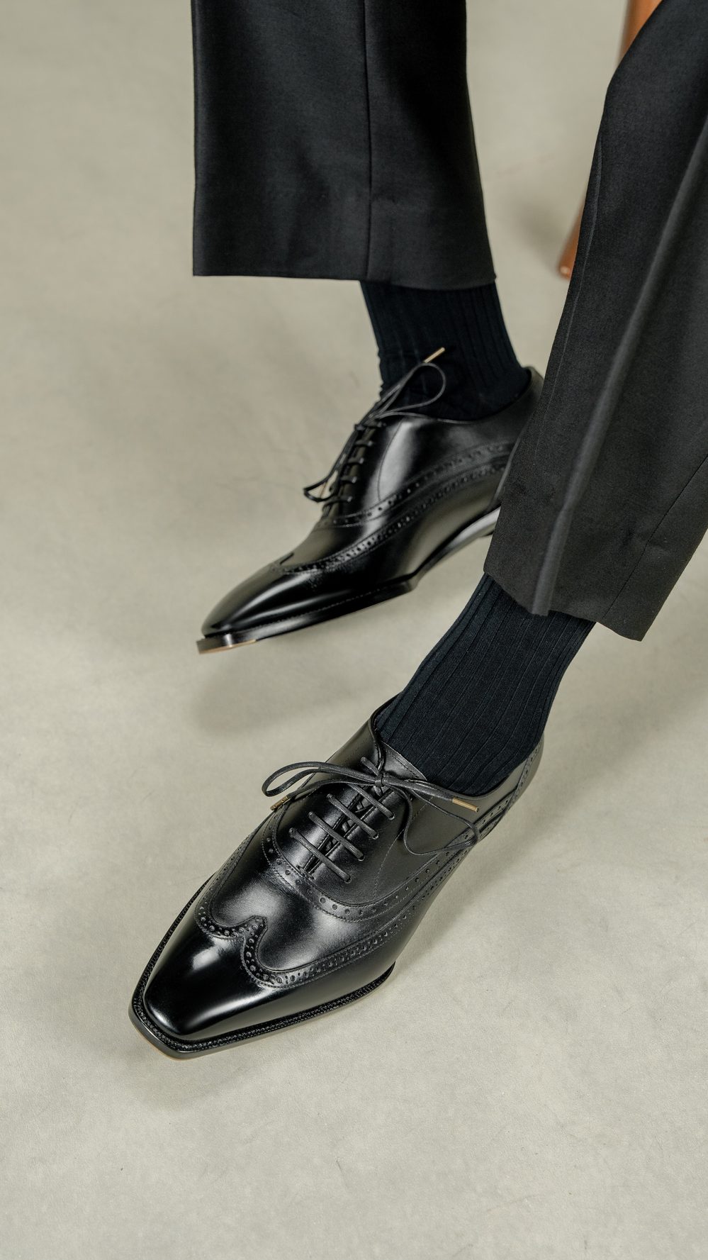 A pair of black leather dress shoes with intricate detailing, worn with dark ribbed socks.