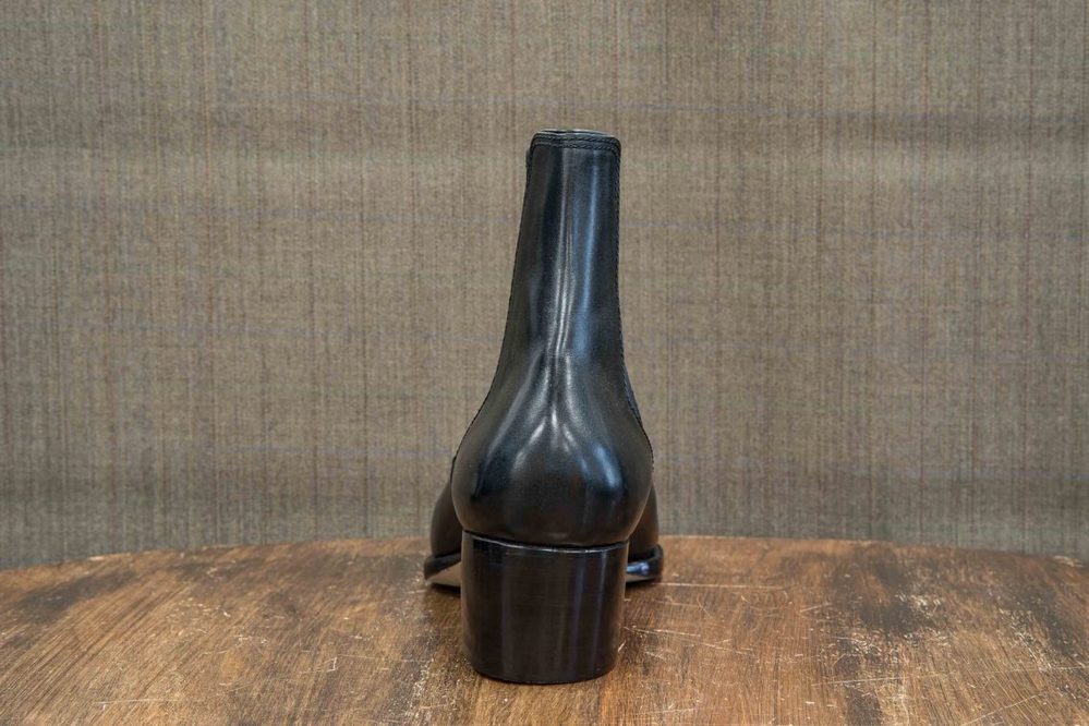A sleek black high-heeled boot is displayed from the back, showcasing its smooth leather material and rounded heel.