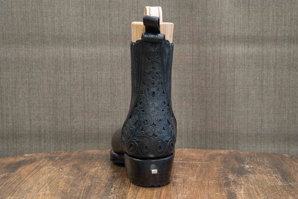 A black leather boot with intricate embossed patterns and a wooden last visible at the top.