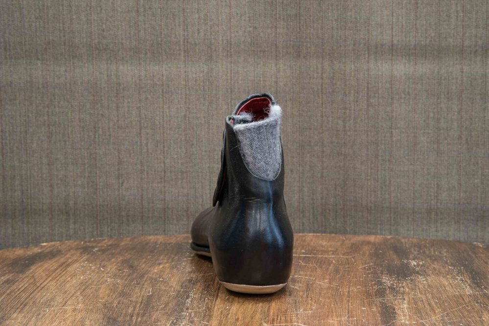 A black leather boot with a gray felt collar and red lining, viewed from the back.