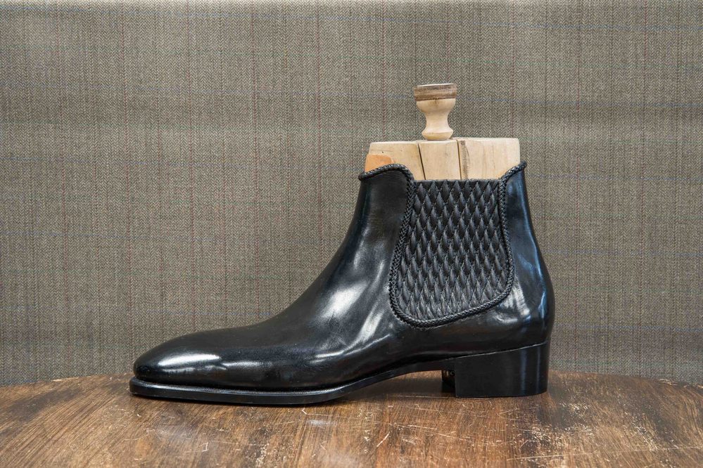 A polished black leather ankle boot with a woven texture on the side and a wooden shoe tree inserted.