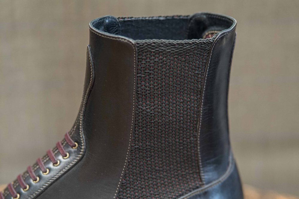 A close-up of a dark brown leather boot featuring a woven textile panel and burgundy laces.