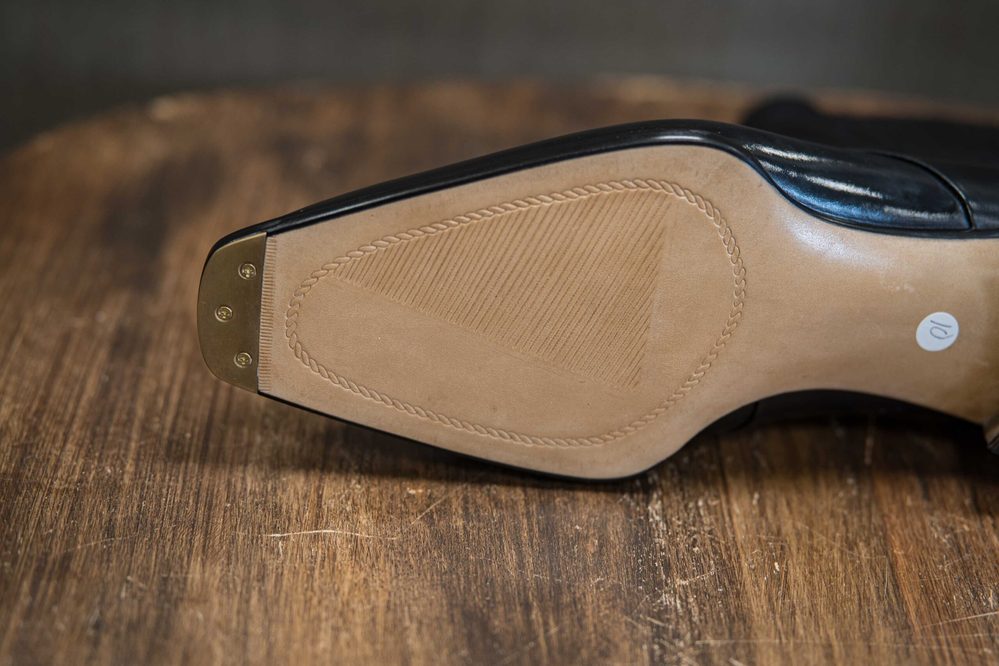 Close-up of the sole of a black shoe featuring a smooth tan leather surface with a textured pattern and gold metal details.