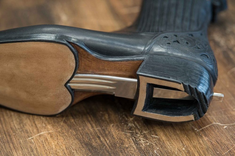 A close-up of a black leather boot sole featuring a unique metallic heel design and a smooth, tan leather bottom.
