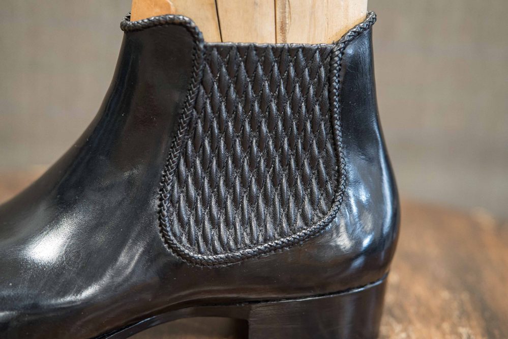 A close-up of a black leather ankle boot featuring a textured woven panel on the side.