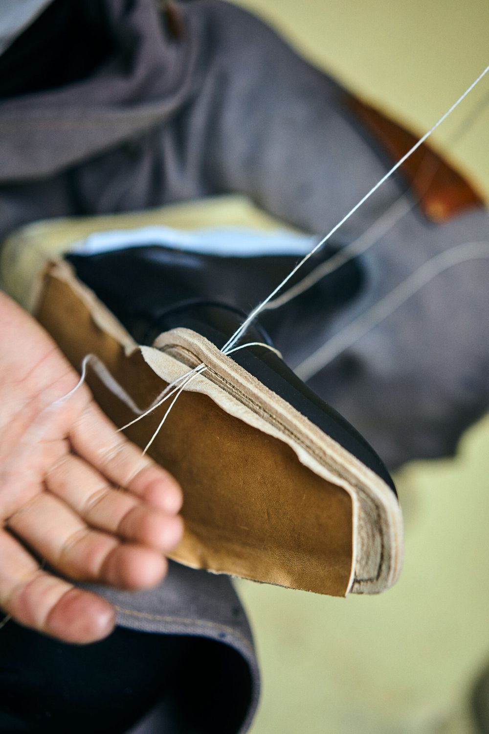 A hand holds a leather shoe with a visible tan sole, as white thread is being stitched through it.