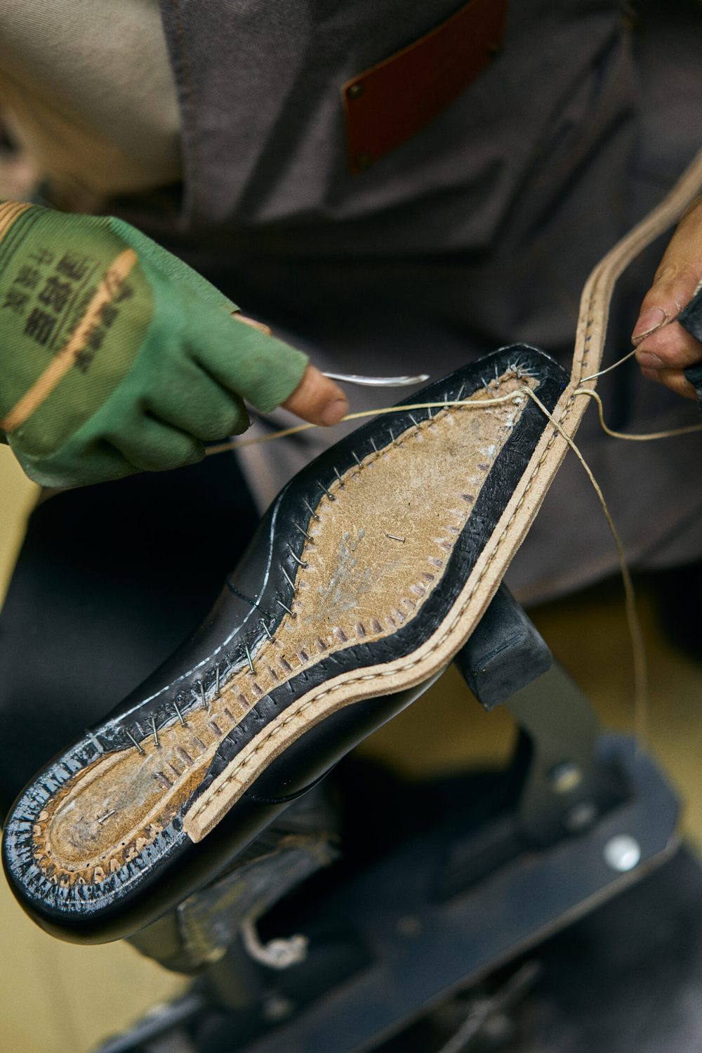 A person with green gloves stitches a black shoe sole with beige thread.