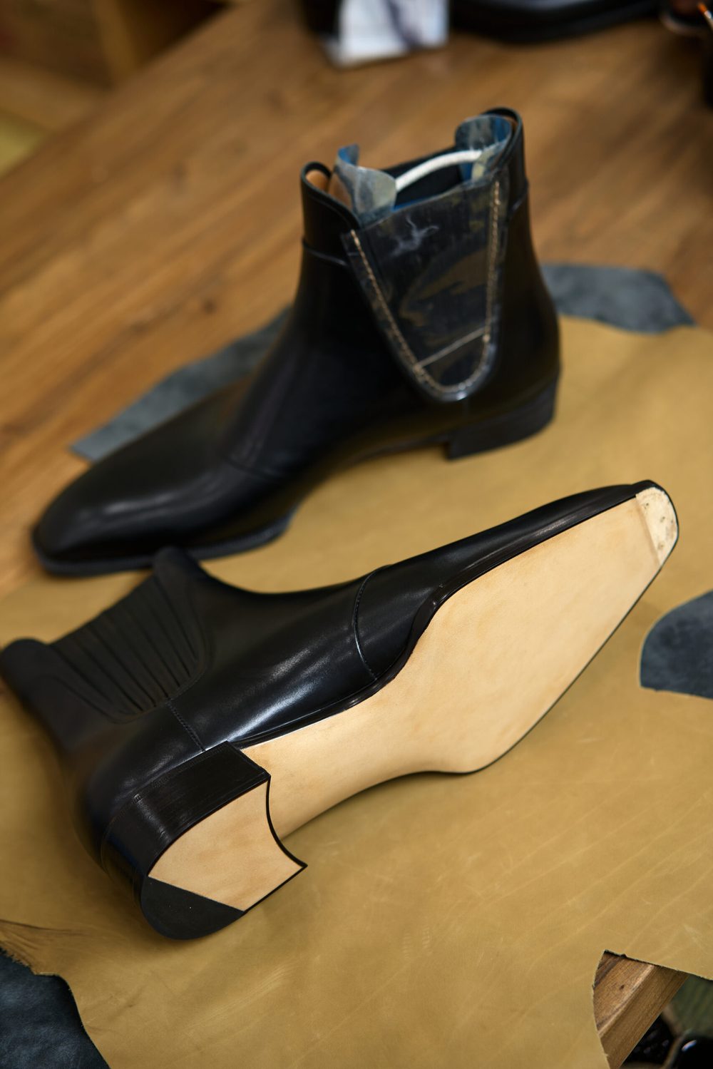 A pair of sleek black leather boots with a tan sole, featuring a distinctive design and textured paneling.