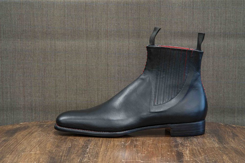 A sleek black leather ankle boot featuring red stitching and elastic side panels.