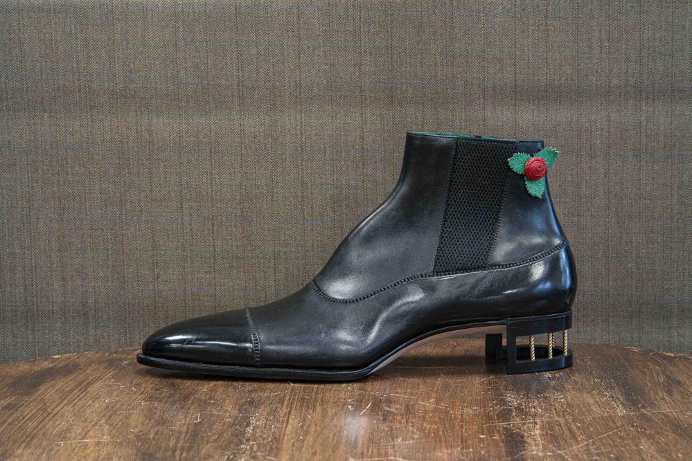 A black leather ankle boot with a decorative rose and green leaves on the side and a unique block heel.