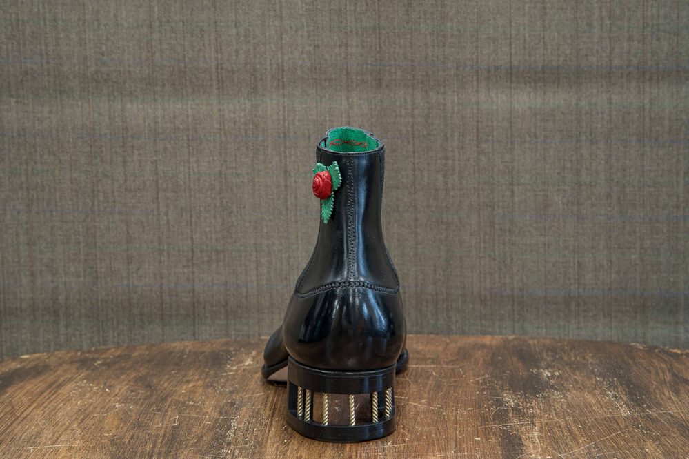 A black leather ankle boot with a decorative red rose and green leaves on the back.