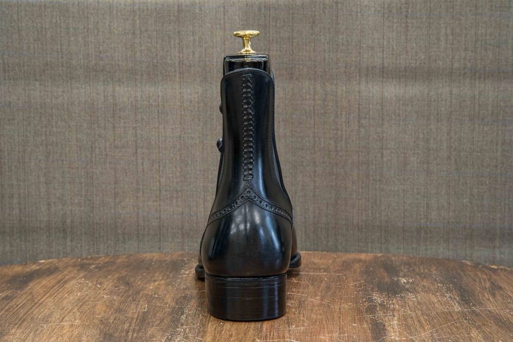 A black leather boot with decorative stitching and a gold heel lift is shown from the back.