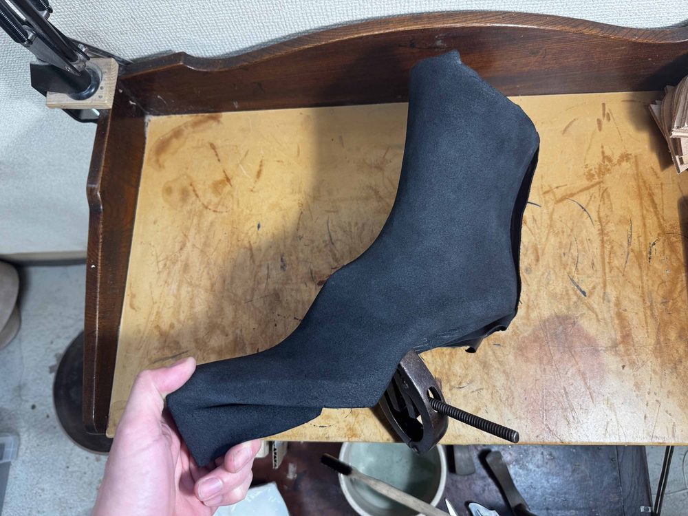 A hand holds a black suede shoe upper on a wooden workbench.
