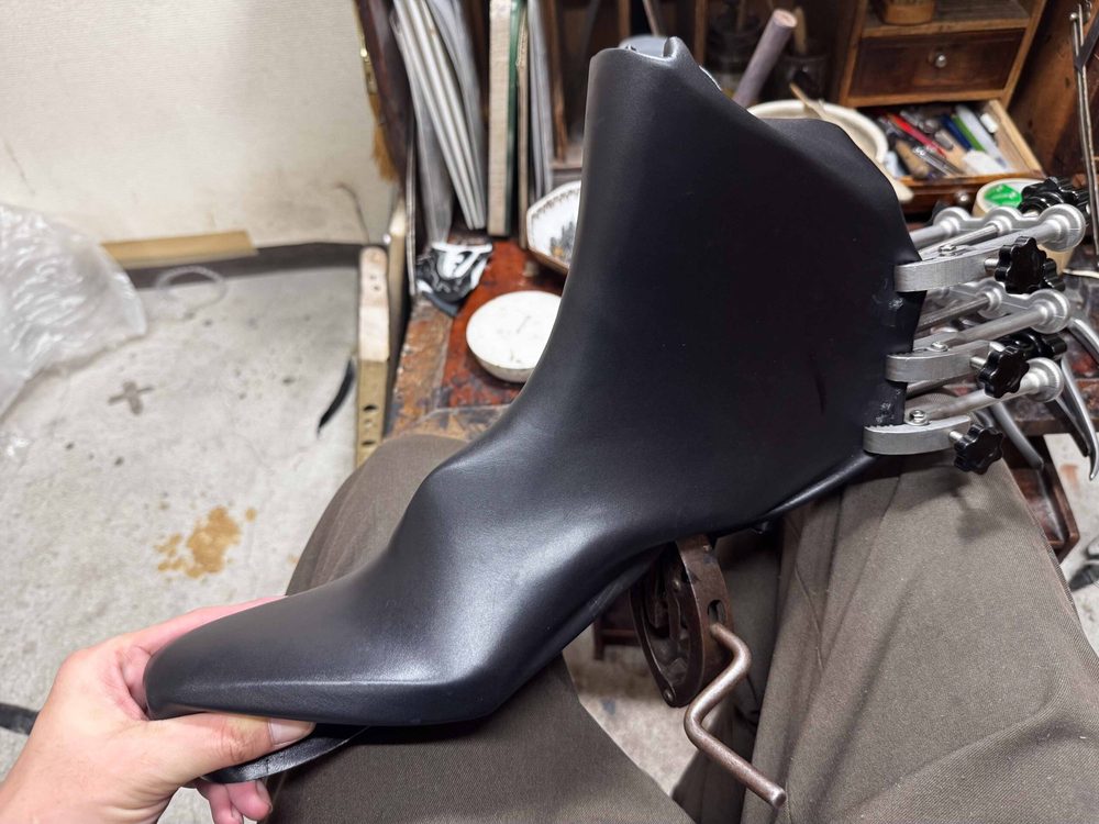 A black shoemaking mold is held in a hand, showing its curved shape and metallic clamps.