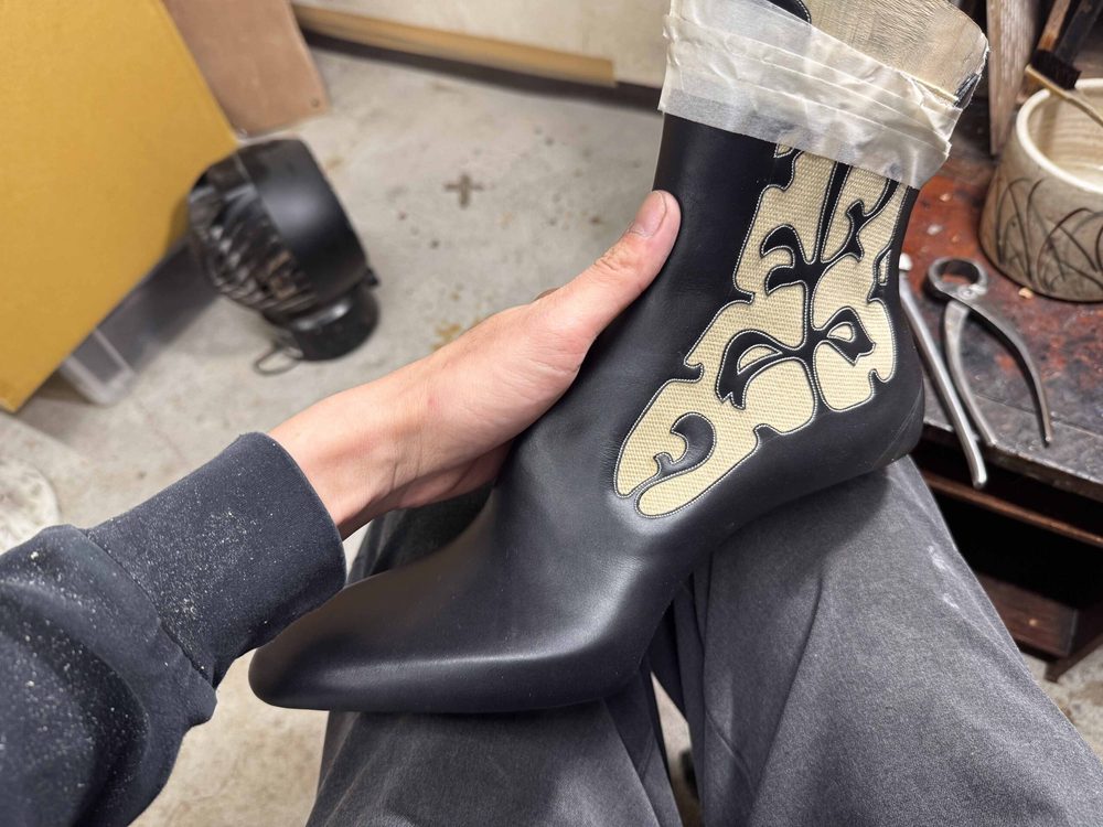 A hand holds a black leather boot with a decorative beige design on the side.