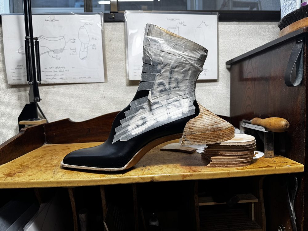 A black leather boot with a wooden heel and tape wrapped around the upper section is displayed on a workbench.