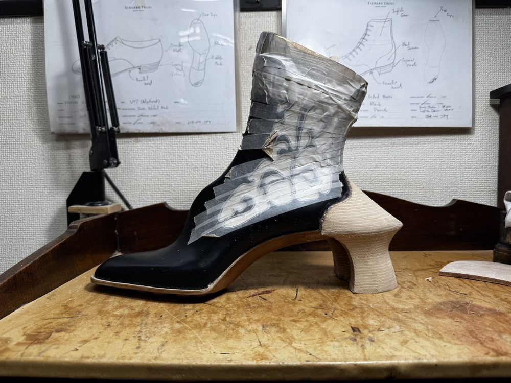 A black shoe prototype with a unique wooden heel, partially covered in tape, is displayed on a wooden surface.