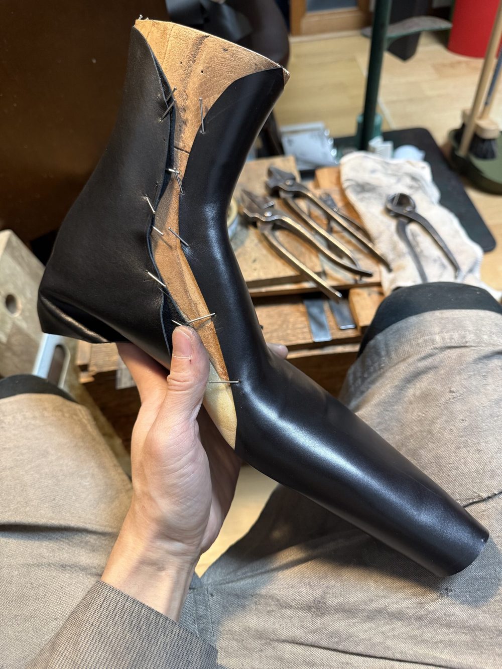A hand holds a partially assembled black leather shoe, showcasing stitching and a wooden sole.
