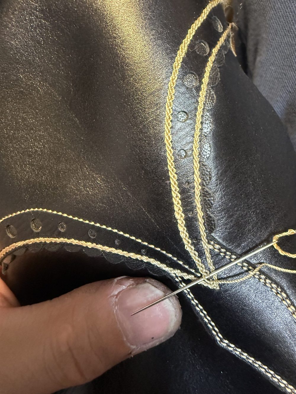 A close-up of a hand holding a sewing needle, stitching golden thread into a black leather shoe.