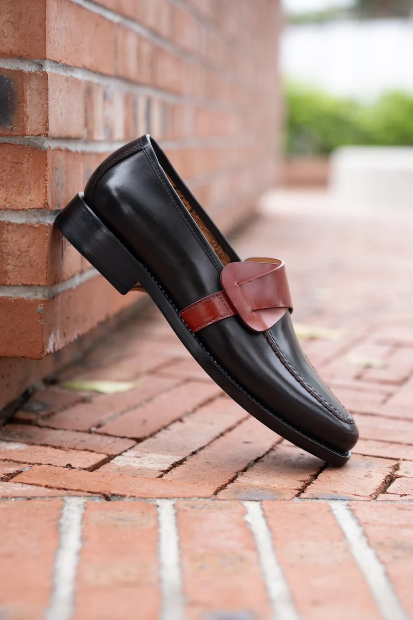 A black leather shoe with a brown leather strap detail, positioned at an angle against a brick wall.