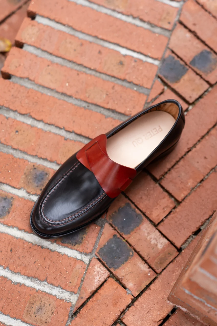 A black leather shoe with a red leather strap across the top.