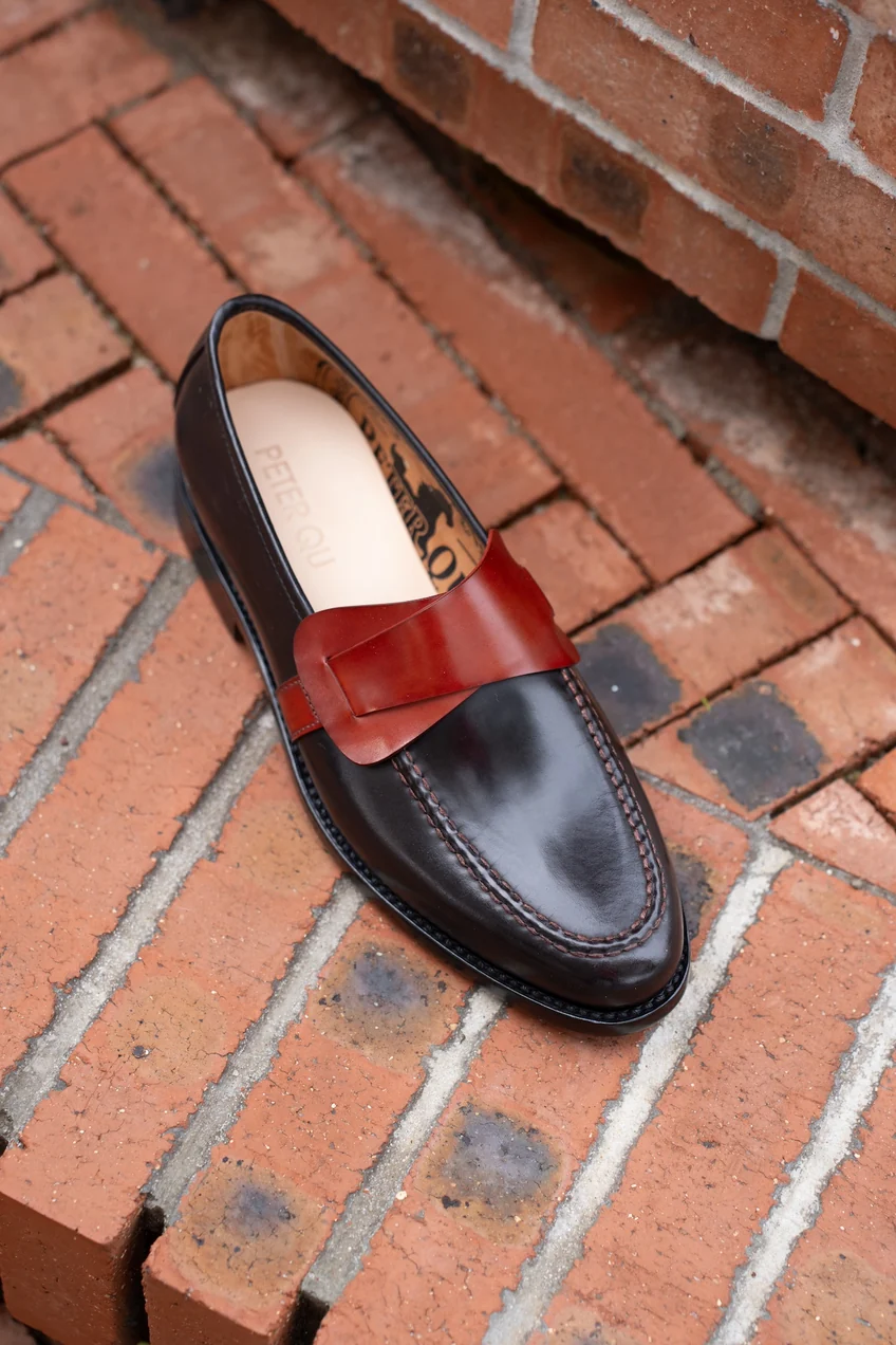 A dark brown leather shoe with a red leather strap across the top, featuring detailed stitching.