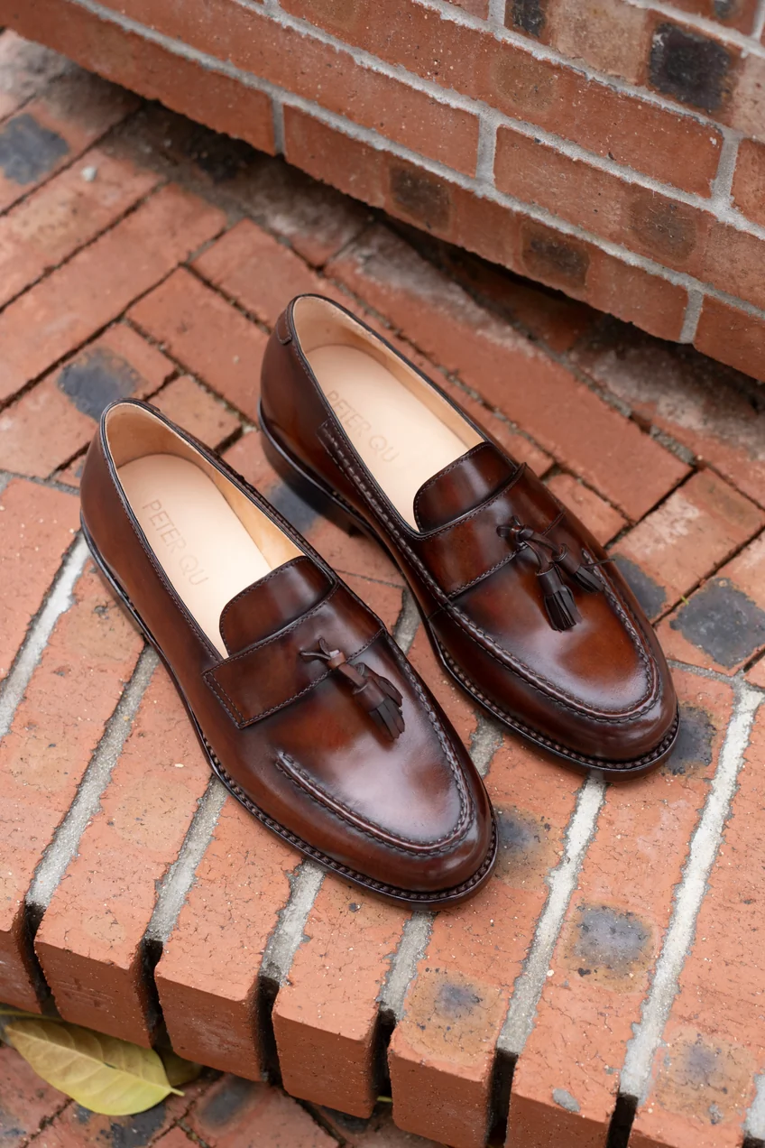 A pair of brown cordovan loafers with tassels and a stitched design.