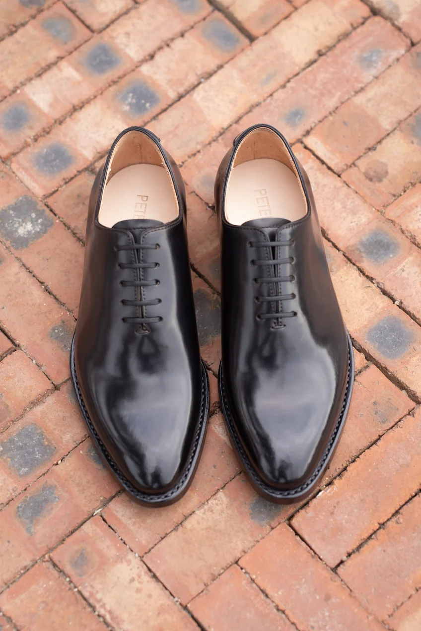 A pair of polished black leather shoes with a pointed toe and six eyelets.